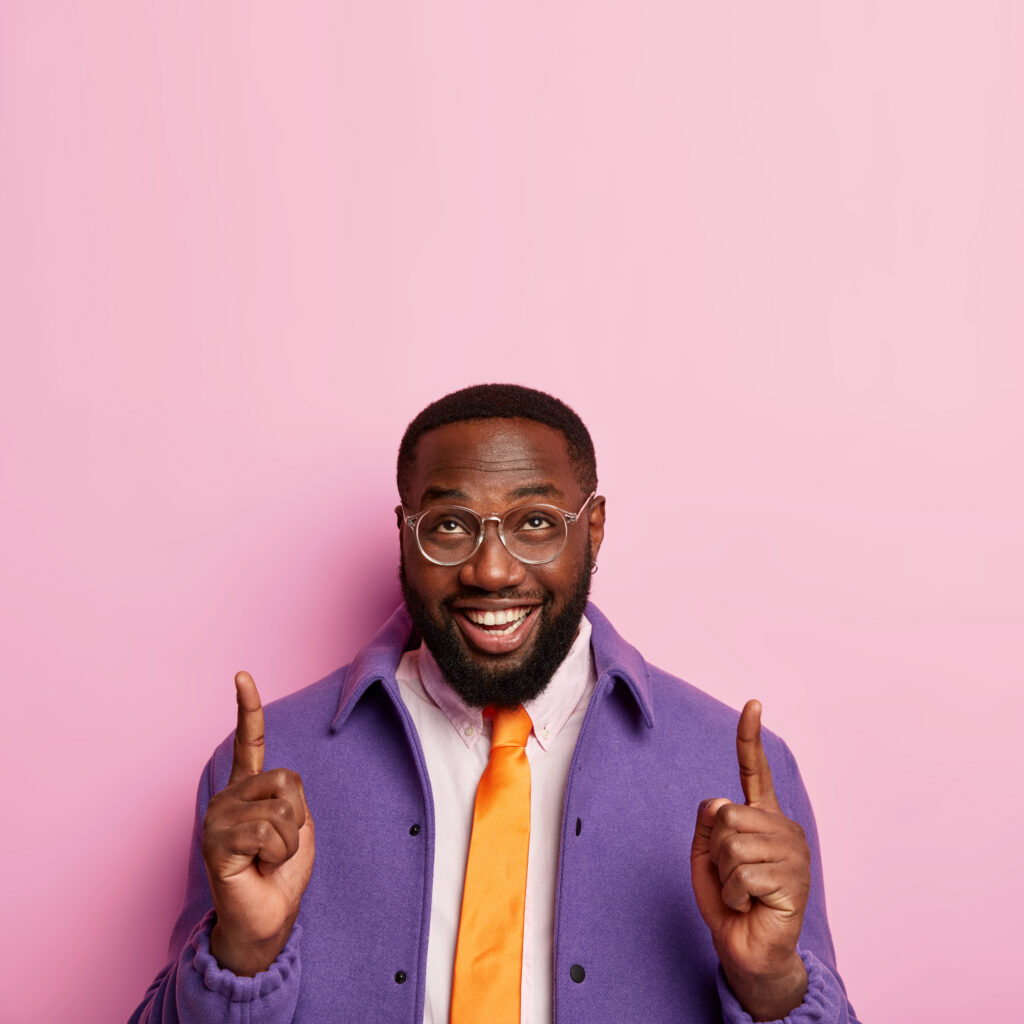 cheerful unshaven man points both index fingers up, demonstrates successful idea, promots copy space, wears spectacles, orange tie and purple coat, isolated over rosy background, introduces promo